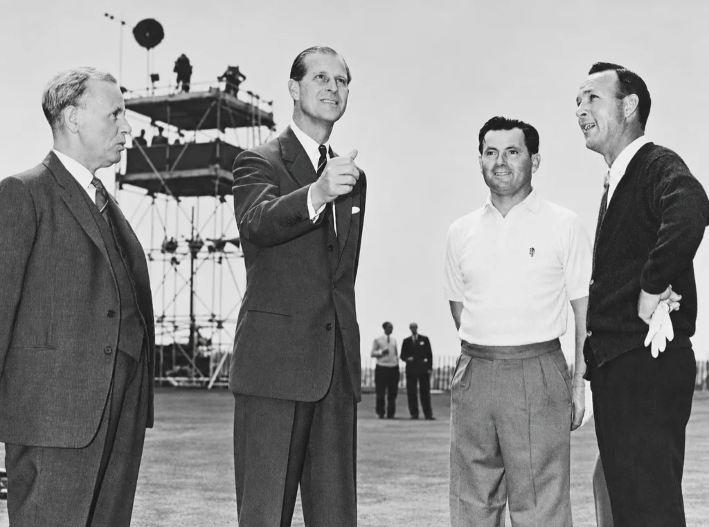 Black and white photograph from 5 July 1963: Prince Philip gestures as he tosses the coin on the 18th green, Club Captain Norman Lees on his right and the two captains, Dai Rees and Arnold Palmer, on his left, with a tall scaffold tower behind them.